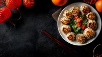 Steamed dumplings with meat filling garnished with fresh herbs and chili on white plate, served with chopsticks. Traditional Asian cuisine on dark background with red lanterns.