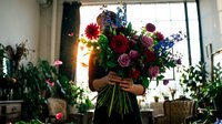 Florist holding vibrant mixed flower bouquet with red, pink, and blue blooms in bright studio setting with potted plants and natural window light for floral business and arrangement concepts.