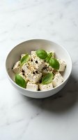 Fresh feta cheese cubes with basil leaves and black pepper in white bowl on marble surface for Mediterranean cuisine and healthy eating.