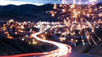 Vibrant fireworks burst over winding mountain road with long exposure light trails and city lights glowing in valley below during nighttime celebration.
