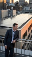 Caucasian male professional in dark blazer and light blue shirt standing on urban rooftop terrace at sunset, overlooking city skyline with modern buildings, suitable for business, corporate, and lifestyle concepts.