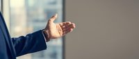 Businessman in dark suit extending open hand in welcoming gesture against blurred office window background, representing trust, partnership, and professional communication.
