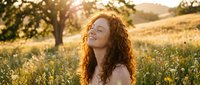 Young woman with curly red hair enjoying peaceful moment in golden hour sunlight among wildflowers in countryside meadow with trees in background.