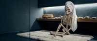 Skeleton wearing white cloth sitting on sand in dark museum setting with ancient artifacts and bones displayed on shelf behind.