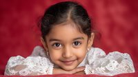 Young Hispanic girl with dark hair in white lace dress smiling at camera against red bokeh background for portrait photography and family concepts.