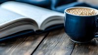 Fresh coffee in navy blue cup with foam next to open book on rustic wooden table, morning reading atmosphere with soft natural lighting.