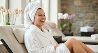 Happy Asian woman in white bathrobe and towel relaxing on sofa after spa treatment at luxury wellness center with flowers and candles in background.