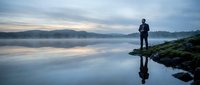 Man standing by calm lake at dawn with mountain reflection in misty water creating peaceful outdoor meditation scene for wellness and mindfulness concepts.