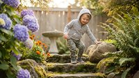 Cheerful toddler boy in striped hoodie exploring a lush garden with hydrangeas, stone steps, and birdbath in warm natural light.