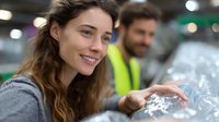 Young woman with long wavy brown hair smiling while recycling plastic bottles at collection center, with worker in safety vest visible in background.