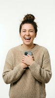 Young woman with brown hair in top knot bun, Caucasian ethnicity, wearing beige knit sweater, holding smartphone to chest, laughing with genuine joy and excitement against white background.