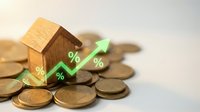Wooden house model with green percentage arrow showing mortgage rate growth surrounded by golden coins on white background for financial planning.