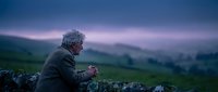Senior Caucasian man in warm clothing sitting on stone wall during twilight hour, contemplating peaceful countryside landscape with rolling hills.