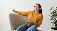 Young Asian woman in yellow sweater laughing joyfully while listening to music with headphones, sitting on beige sofa in modern living room interior.