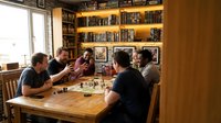 Group of diverse male friends playing board game together at wooden table in cozy home library with bookshelves during casual social gathering.