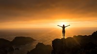 Person with raised arms celebrating success on rocky cliff overlooking ocean at golden sunset. Motivational achievement concept for wellness and personal growth.