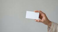 Person of medium skin tone holding blank white business card or paper against neutral gray background, suitable for corporate branding and marketing materials.