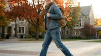 Young male student with brown hair and light skin tone wearing denim jacket and jeans, carrying backpack, walking across campus courtyard during autumn with fall foliage and historic stone buildings.