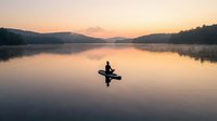 Silhouette of person kayaking on calm lake at sunrise with mountains and forest reflection in still water creating peaceful morning scene.