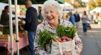 Happy senior Caucasian woman with white curly hair holding fresh vegetables in reusable bag at farmers market, smiling joyfully while shopping for organic produce.