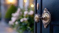 Ornate brass door handle on navy blue door with blurred pink flowers and bokeh lights in background creates welcoming entrance atmosphere.