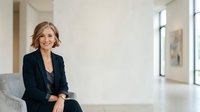Middle-aged woman with blonde wavy hair, Caucasian ethnicity, wearing dark blazer and black pants, sitting confidently on gray sofa in modern bright office or home interior with natural light.