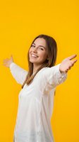 Young woman with long brown hair and fair complexion wearing white casual shirt, smiling with arms spread wide against bright yellow background for lifestyle and happiness concepts.