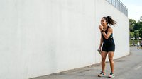 Young woman with dark hair and medium skin tone wearing black athletic wear, stretching her arms during outdoor fitness routine against white concrete wall.