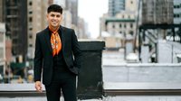 Young Hispanic businessman in black suit with orange shirt walking confidently through modern urban city street with skyscrapers in background.