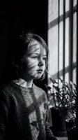 Young girl in knitted sweater standing by window with dramatic light and shadow patterns in black and white monochrome portrait for emotional storytelling.