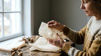 Middle-aged Caucasian woman in cozy sweater reading old letters by window with autumn decorations creating nostalgic atmosphere for lifestyle content.