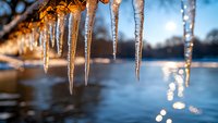 Glowing icicles hanging against blurred winter lake background with golden sunset light and bokeh reflections creating magical winter atmosphere.