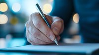 Close-up view of hand holding metal pen while writing on paper document against blurred bokeh lights background, capturing professional business moment.