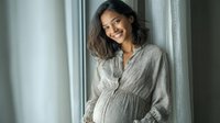 Young pregnant woman with long dark hair smiling by window in soft gray maternity dress, radiating warmth and joy during pregnancy.