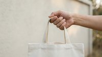 Person holding white cotton tote bag with handles for shopping, retail, branding, or promotional use.