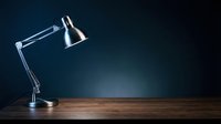 Adjustable metal desk lamp with chrome finish positioned on wooden surface against dark blue background, suitable for office and workspace design projects.