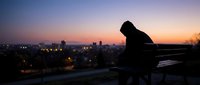 Silhouette of contemplative man sitting alone on bench overlooking city lights at sunset, expressing solitude, depression, and mental health struggles.