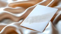 Blank white paper card on elegant beige silk fabric with soft folds and waves creating luxurious backdrop for wedding invitations or elegant announcements.