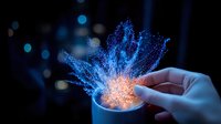 Glowing magical particles burst from cup in dark setting, with hand touching ethereal blue and orange light explosion effect against black background.