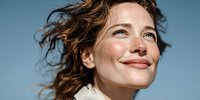 Young woman with freckles and curly hair smiling against blue sky, expressing happiness and optimism in natural sunlight.