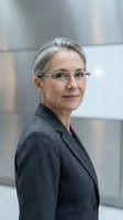 Mature woman with gray hair wearing eyeglasses and dark blazer, smiling at camera against modern office background. Corporate portrait for business, professional profiles, leadership representation.