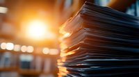 Stack of paper documents with warm sunset light through office window creating atmospheric bokeh effect and dramatic shadows on business paperwork files.