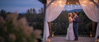 Romantic wedding couple dancing under elegant white curtained gazebo at twilight with string lights and mountain landscape background.