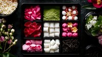 Decorative box with rose petals, matcha powder, lychee fruits and sugar cubes arranged in compartments on dark background, styled for Asian tea ceremony.