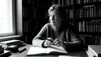 Elderly Caucasian man writing in notebook at desk surrounded by books in home library. Black and white portrait of senior author or scholar working on manuscript.