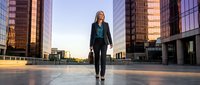 Professional businesswoman walking confidently in modern urban plaza at sunset with glass skyscrapers and corporate buildings creating dynamic cityscape backdrop.