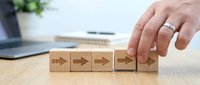 Male hand placing wooden blocks with arrow symbols showing business process steps and workflow progression on office desk.