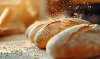 Fresh baked bread loaves dusted with flour on wooden surface, captured in warm lighting with floating flour particles creating magical bakery atmosphere.