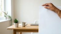 Male hand holding blank white paper sheet against neutral interior background with wooden desk, potted plant, and cup for mockup and branding purposes.