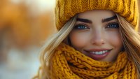 Young Caucasian woman with bright blue eyes wearing yellow knitted beanie and scarf smiling warmly on blurred autumn background, close up portrait.